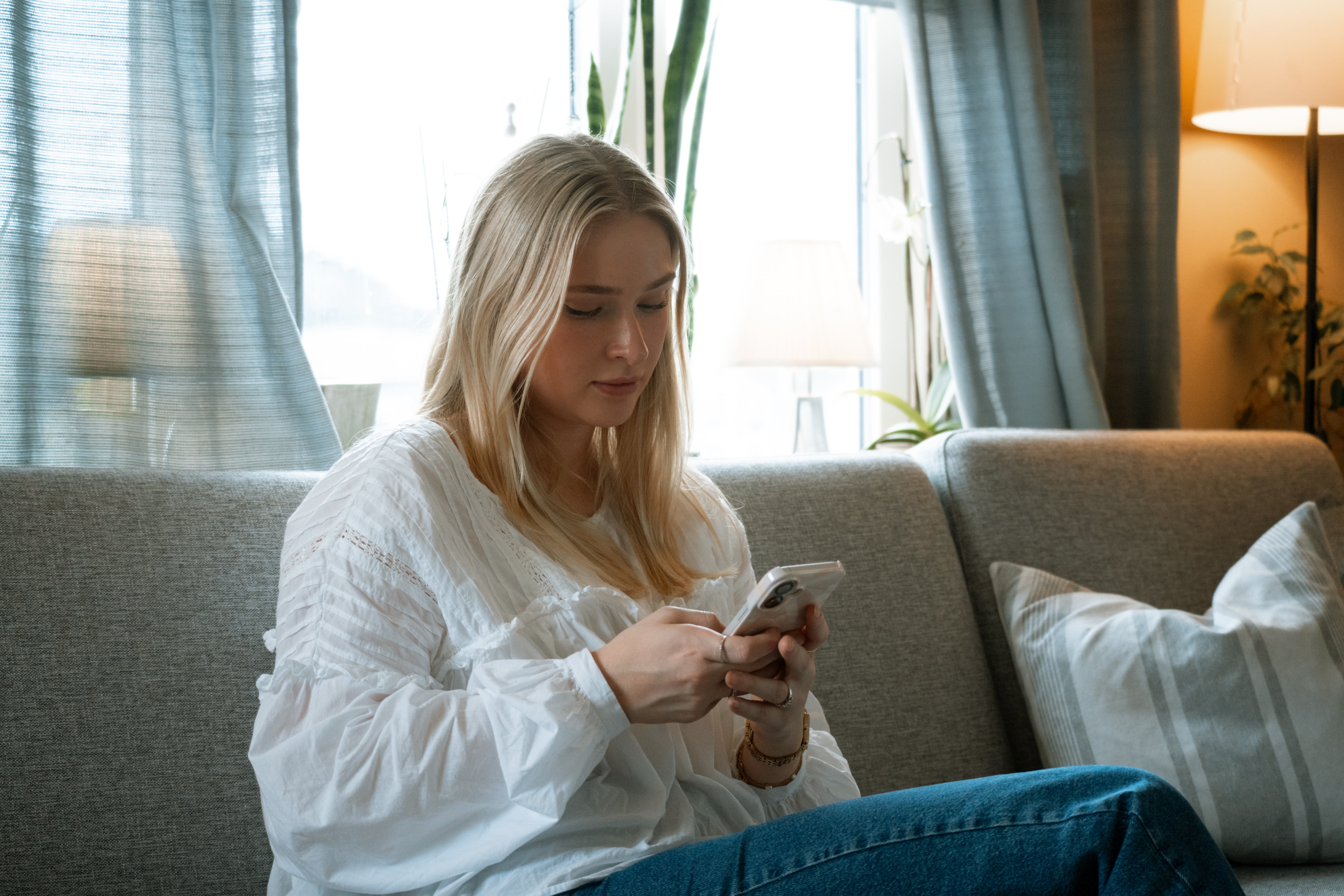 Person med hvit bluse sitter i sofaen og holder en mobil, med klokke på håndleddet.