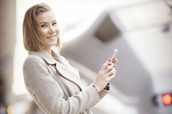 Smilende kvinne holder mobilen sin. Det er et tog i bakgrunnen.